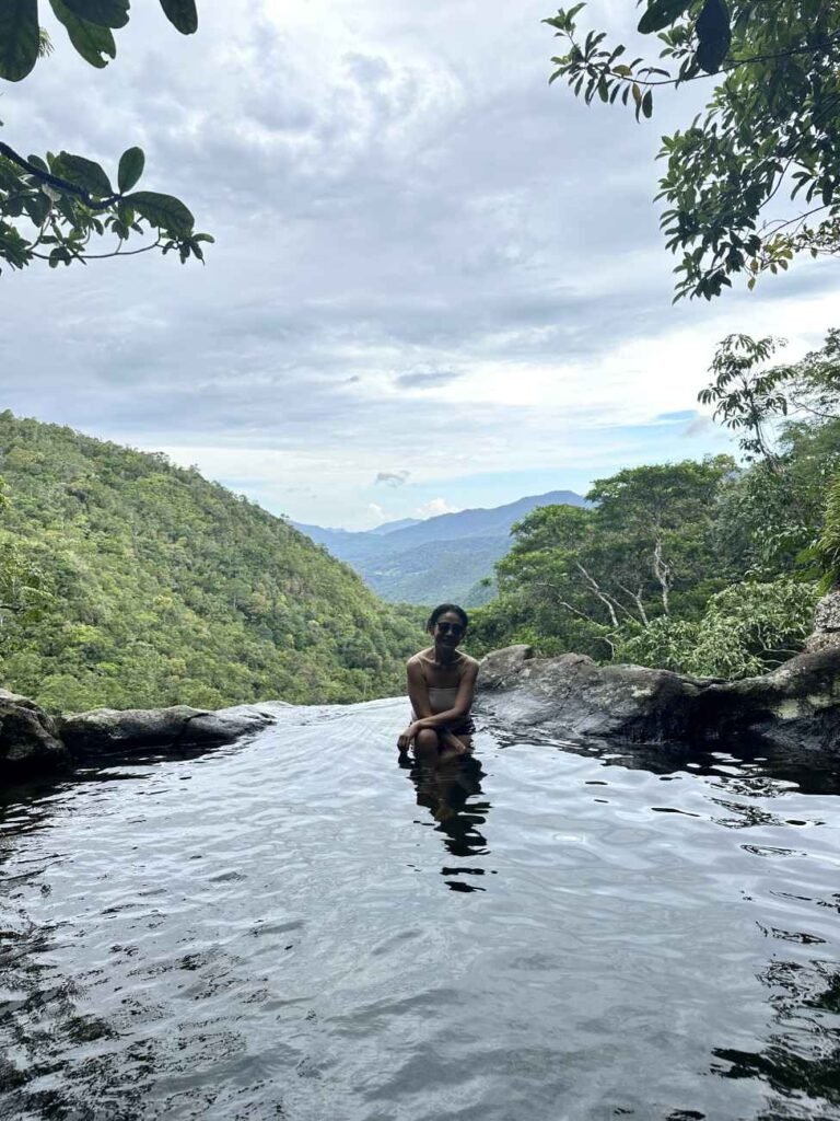 Woman standing in a river at Cunca Pilas Waterfall surrounded by lush greenery and mountainous landscape.