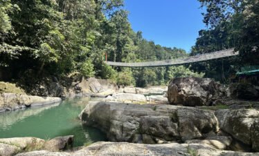 Person jumping from rock into the crystal-clear water at Cunca Wulang Waterfall with bamboo bridges in the background.