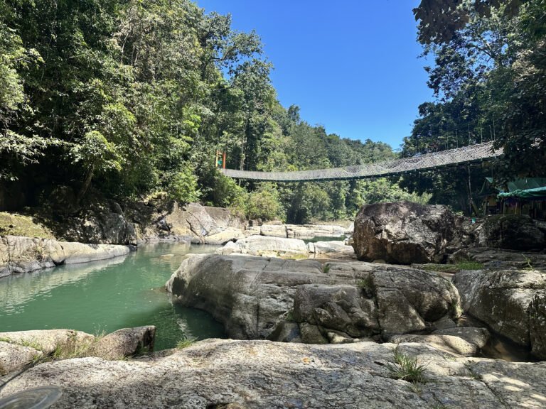 Person jumping from rock into the crystal-clear water at Cunca Wulang Waterfall with bamboo bridges in the background.