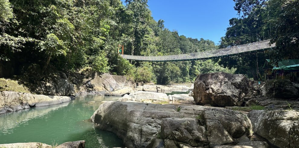 Person jumping from rock into the crystal-clear water at Cunca Wulang Waterfall with bamboo bridges in the background.