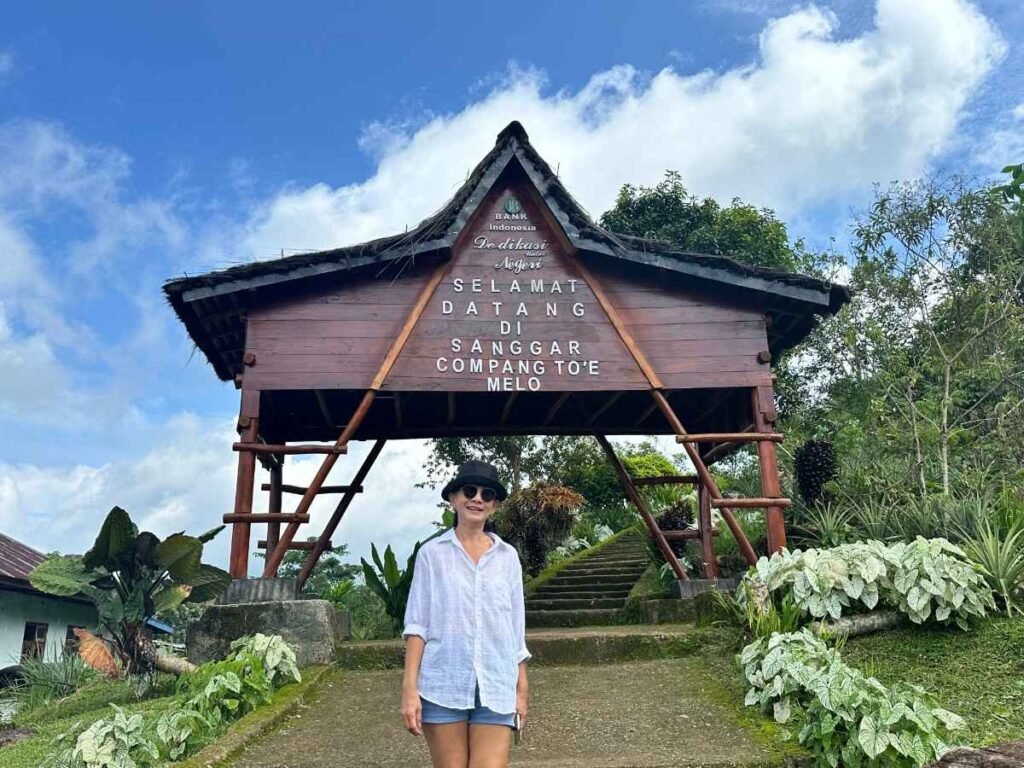 A Local’s Guide to Hidden Indonesia Adventures 6 A woman standing in front of the entrance sign to Sanggar, with a traditional house and lush greenery around.Flores Indonesia cultural tours