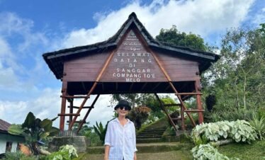 A woman standing in front of the entrance sign to Sanggar, with a traditional house and lush greenery around.Flores Indonesia cultural tours