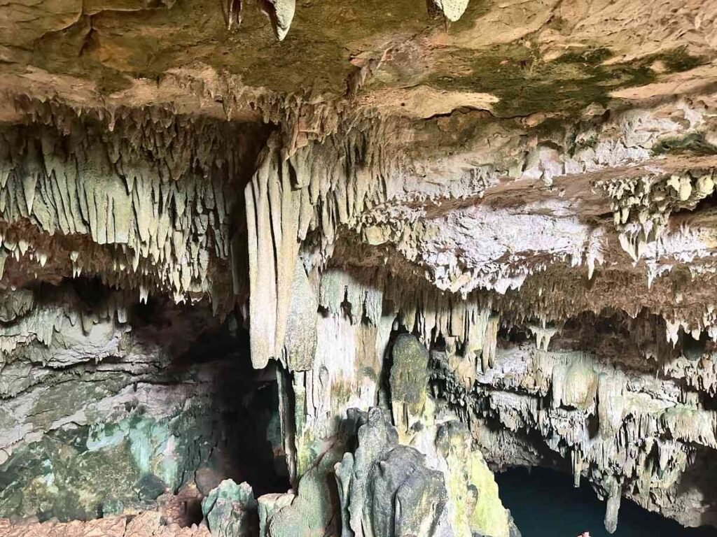 Inside a large cave with stalactites hanging from the ceiling and a vibrant, clear blue water pool filled with swimmers.