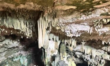 Inside a large cave with stalactites hanging from the ceiling and a vibrant, clear blue water pool filled with swimmers.