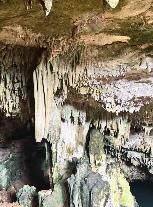 Inside a large cave with stalactites hanging from the ceiling and a vibrant, clear blue water pool filled with swimmers.