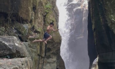 Water cascading down a high rocky cliff into a pool below with greenery on both sides.cunca wulang