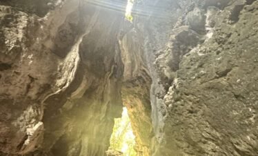 Inside a cave with sunlight streaming through a hole above, illuminating the rocky interior.