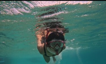 Person snorkeling underwater with goggles and snorkel, surrounded by blue water and marine life.