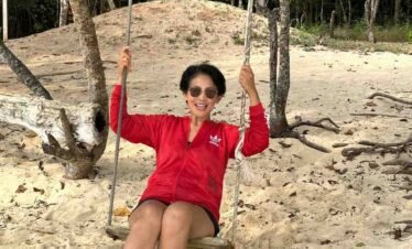 Woman in sunglasses smiling and sitting on a wooden swing hung from trees on a sandy beach.